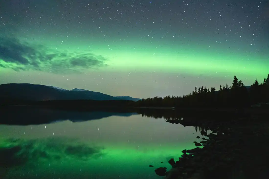 Green aurora borealis over a still lake, mirrored in the water, with a forested shoreline and low mountains under a starry sky.