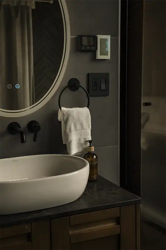 Dimly lit bathroom with a round backlit mirror, white basin on a dark stone counter, black fixtures and a white hand towel.