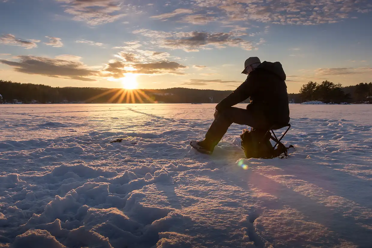Person sitting on a folding stool on a frozen snow-covered lake, facing the setting sun