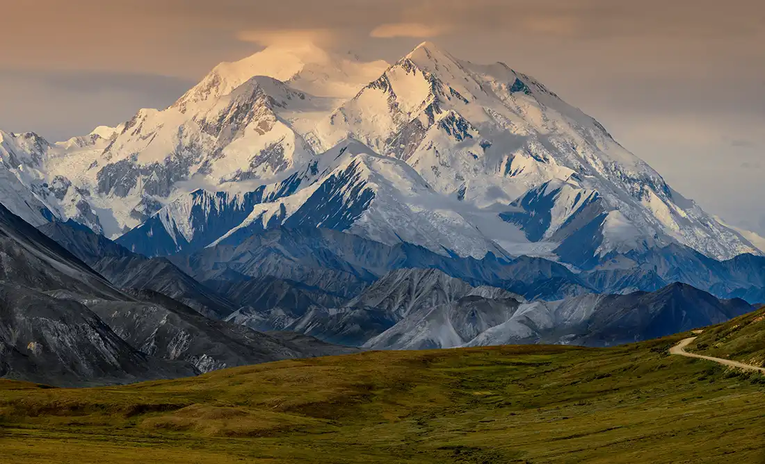 Large snow-covered mountain massif rising above layered ridges and rolling green tundra under a hazy sky.