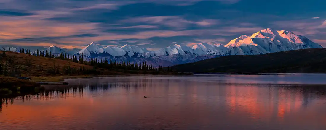 Panoramic snow-capped mountain range lit by sunset, reflected in a calm lake with autumn tundra and spruce along the shore.