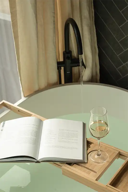 Open book and a glass of white wine on a wooden bath tray across a filling soaking tub with a running black faucet.