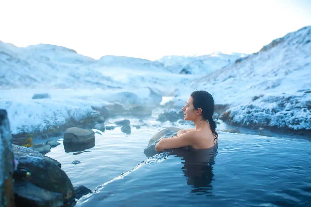 Woman resting at the edge of a steaming hot spring surrounded by snow-covered rocks and hills