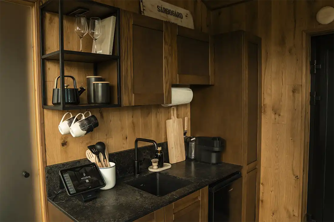 Wood-paneled kitchenette with a black stone counter, sink, open shelving with a kettle and glasses, hanging mugs and a tablet.